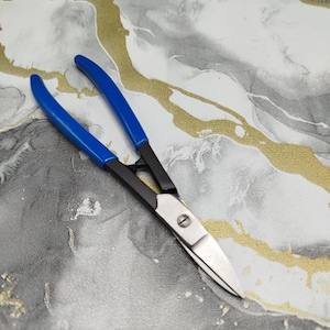 May include: A pair of metal snips featuring bright blue rubberized handles, a black metal frame, and sharp silver-colored steel blades. The tool is designed with a central pivot screw and a spring mechanism between the handles for operation.
