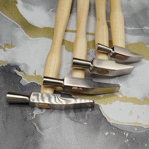 Op de afbeelding: Een set van vier metalen hamers met lichtgekleurde houten handvatten, gerangschikt in een waaierpatroon. Elke hamer heeft een kop van gepolijst staal met een wigvormige punt en een kleine, ronde, messingkleurige inleg aan de zijkant.