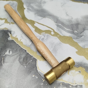 May include: A mallet featuring a light-colored wooden handle and a cylindrical brass head. The brass head consists of two smooth, flat ends and a central section with a textured, cross-hatched knurled pattern for improved grip during use.