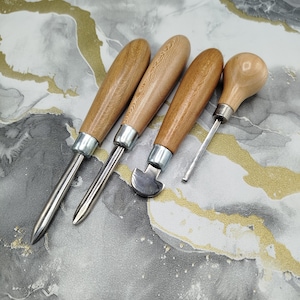 May include: A set of four leatherworking tools featuring smooth, light-colored wooden handles and silver-toned metal ferrules. The tools include two pointed awls, one curved edge creaser, and one small, thin metal punch with a rounded wooden handle.