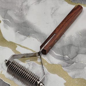 Op de afbeelding: Een verzorgingsinstrument voor huisdieren met een lange, donkerbruine houten handgreep bevestigd aan een roestvrijstalen frame met een reeks scherpe, gekartelde metalen tanden voor het ontklitten en uitdunnen van de vacht.