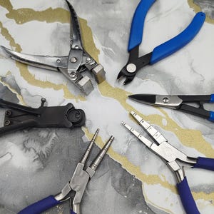 Op de afbeelding: Een verzameling van vijf metalen tangen en knippers voor het maken van sieraden, gerangschikt in een cirkel. De set bevat zilverkleurige platbektangen, zijkniptangen met blauwe handvatten, zwarte tangen en twee rondbektangen met blauwe handvatten.