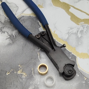 Può includere: Un utensile manuale in metallo nero con impugnature in gomma blu, dotato di un meccanismo a testa circolare con regolazione a vite. Sotto l'utensile si trovano due piccoli anelli, uno in ottone metallico e l'altro in plastica bianca liscia.