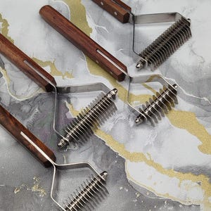 Op de afbeelding: Drie vachtkammen voor huisdieren, elk met een lange, donkerbruine houten handgreep bevestigd met twee metalen klinknagels. De kop van elk gereedschap bestaat uit een roestvrijstalen frame met scherpe, dunne, parallelle metalen tanden.
