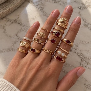 May include: A collection of gold-toned rings worn on fingers, featuring various designs including textured bands, smooth bands with rectangular red stones, a heart-shaped red stone, small white pearls, and chain-link patterns in a stacked arrangement.