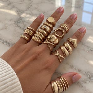 Può includere: Una collezione di anelli color oro indossati su tutte le dita, incluso il pollice. Il set presenta vari design come fasce lisce, texture intrecciate, un anello con una piccola pietra chiara, un anello aperto circolare e fasce metalliche incrociate.