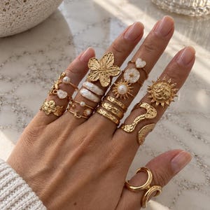 Puede incluir: Una colección de anillos dorados usados en los dedos, con varios diseños que incluyen una flor, un sol, un corazón, motivos celestiales, perlas y bandas texturizadas. Los anillos varían en forma y tamaño, mostrando acabados de metal pulido.