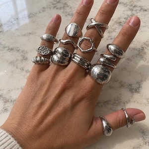 May include: A collection of fifteen silver-toned rings worn across all fingers and the thumb. The rings feature various designs including smooth bands, textured surfaces, organic shapes, a cross-over style, and a large rounded dome ring.