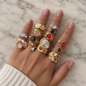 May include: A collection of gold-toned rings worn on fingers, featuring various designs including textured bands, star shapes, and settings with iridescent, red, brown, and clear stones. The rings display diverse shapes, from smooth cabochons to sunburst motifs.