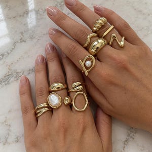 Puede incluir: Una colección de anillos dorados usados en los dedos, con varios diseños que incluyen bandas lisas, superficies texturizadas, un anillo con una perla blanca, un anillo con una piedra blanca ovalada y formas metálicas abstractas y onduladas.
