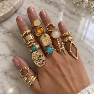 Op de afbeelding: Een collectie goudkleurige ringen gedragen aan de vingers, met diverse ontwerpen waaronder gladde banden, getextureerde oppervlakken, geometrische vormen en ringen bezet met amberkleurige, turquoise en witte stenen in diverse stijlen.