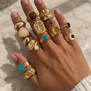 Op de afbeelding: Een verzameling goudkleurige ringen gedragen aan de vingers, met diverse ontwerpen waaronder gladde banden, getextureerde oppervlakken en zettingen met tijgeroog, turkoois, witte, gele en donkerbruine stenen en een zonmotief.