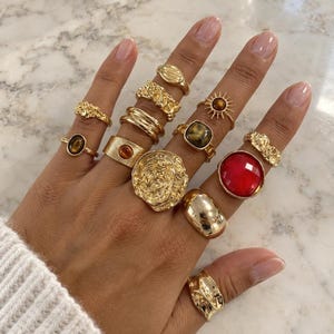 Op de afbeelding: Een verzameling goudkleurige ringen gedragen aan de vingers, met diverse ontwerpen waaronder een grote cirkelvormige ring met textuur, een ring met een gefacetteerde rode steen, ringen met bruine ovalen stenen, een zonnestraalontwerp en diverse ringbanden.