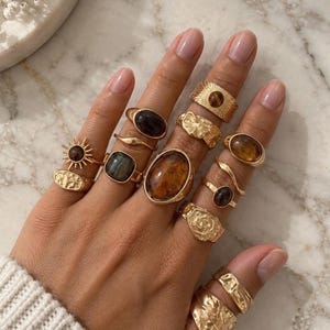 Op de afbeelding: Een verzameling goudkleurige ringen gedragen aan de vingers, met diverse ontwerpen waaronder gladde banden, getextureerde oppervlakken en zettingen met bruine, amberkleurige en donkere stenen. Sommige ringen hebben zonnestraalpatronen en organische vormen.