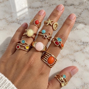 Könnte beinhalten: Eine Sammlung goldfarbener Ringe an den Fingern mit verschiedenen Motiven, darunter ein Seestern, eine Muschel, eine Mondsichel und mehrere Ringe mit bunten Cabochon-Steinen in den Farben Rot, Orange, Gelb, Blau und Rosa.