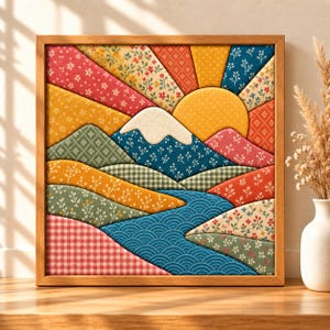 Op de afbeelding: Een vierkant ingelijst kunstwerk aan de muur met een gelaagde stoffencollage van een landschap. De scène toont een berg met een witte top, een grote gele zon en glooiende heuvels in diverse bloemen-, ruit- en geometrische patronen.