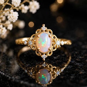 May include: A gold ring featuring an oval-shaped opal center stone with iridescent flashes of green and orange. The setting is crafted with intricate filigree metalwork and accented by four small, clear round-cut diamonds.
