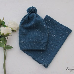 Pode incluir: Um chapéu azul de malha com um pompom no topo e uma echarpe de malha azul combinando. O chapéu e a echarpe têm um padrão de floco de neve.