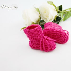May include: A pair of pink knitted baby booties with white laces. The booties are on a white background with white roses.