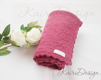 Może przedstawiać: Różowy dzianinowy kocyk dla niemowląt z falbanką. Kocyk jest złożony i ma białą etykietę z napisem "Kairi Design".