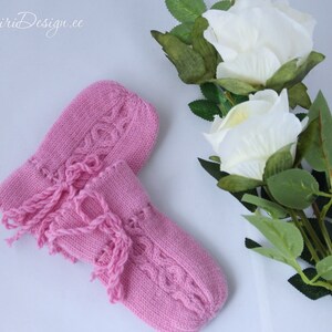 Könnte beinhalten: Ein Paar rosa gestrickte Babyschuhe mit Fransen. Die Schuhe haben ein Zopfmuster und sind aus weichem Garn gefertigt.