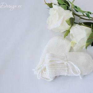 Könnte beinhalten: Ein Paar elfenbeinfarbene Babymützen und -schuhe werden mit weißen Rosen und Grünzeug präsentiert. Die Fäustlinge und Schuhe sind aus weichem Strickmaterial gefertigt. Das Bild hat ein weiches, natürliches Licht.