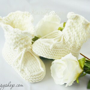 May include: A pair of ivory-colored, knitted baby booties with a textured pattern and delicate ties. The booties are displayed with white roses, creating a soft and gentle aesthetic. The website "katsyshop.com" is visible in the image.