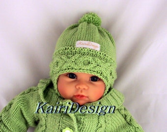 May include: A green knitted baby hat with a pom pom on top and a white label that says "KairiDesign". The hat is worn by a baby doll with blue eyes.