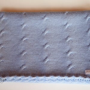 以下が含まれることがあります： 薄い青色のニットのベビーブランケットで、縁はギザギザになっており、小さな白いタグには「Knitted with Love」と書いてあります。