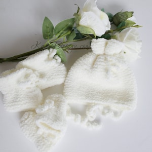 May include: White crocheted baby hat and matching mittens with a fluffy texture. The hat has a pom-pom on top and the mittens have a ruffled edge.