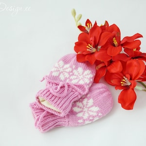 Könnte beinhalten: Ein Paar rosa gestrickte Fäustlinge mit weißen Schneeflockenmustern. Die Fäustlinge sind zusammengefaltet und liegen auf einer weißen Oberfläche neben einem Strauß roter Blumen.