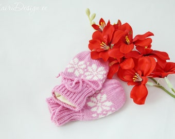 May include: A pair of pink knitted mittens with white snowflake designs. The mittens are folded together and are lying on a white surface next to a bouquet of red flowers.