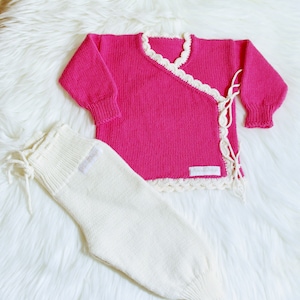 Op de afbeelding: Een roze gebreide babytrui met witte gehaakte rand en een witte gebreide broek.