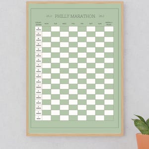 Könnte beinhalten: Ein salbeigrünes Trainingsplan-Poster für den Philly Marathon in einem hellen Holzrahmen. Die Tabelle enthält einen Countdown von 18 bis 1 Woche, mit Spalten für die Wochentage von Montag bis Sonntag sowie eine Spalte für die wöchentlichen Meilen.