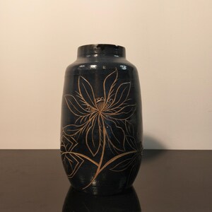 Pode incluir: Um vaso de cerâmica azul-marinho escuro com corpo cilíndrico e gargalo estreito. A superfície apresenta um design floral gravado em cor clara, com uma grande flor em floração exibindo pétalas pontiagudas e folhas num caule central.