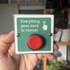May include: A square green sign with a white border featuring the text Everything goes back to normal and a white icon of a hand pressing a button. A large, circular, matte red push button is mounted in the center of the sign with metal screws.