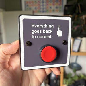 May include: A square wall plate with a dark gray surface, a white border, and a large red circular push button. The text "Everything goes back to normal" is printed in white, accompanied by a white icon of a hand pressing a button.