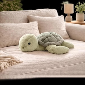 Puede incluir: Un juguete de peluche con forma de tortuga, con pelaje suave de color verde claro y un caparazón texturizado de color verde salvia. El animal de peluche tiene ojos pequeños, oscuros y redondos, y una cabeza redondeada, posado horizontalmente.