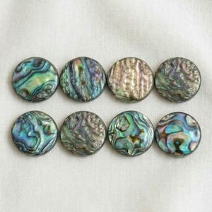 May include: Eight circular buttons crafted from iridescent abalone shell, arranged in two rows of four. Each button displays a unique, swirling pattern of natural colours including deep blues, vibrant greens, soft purples, and shimmering pearlescent tones.