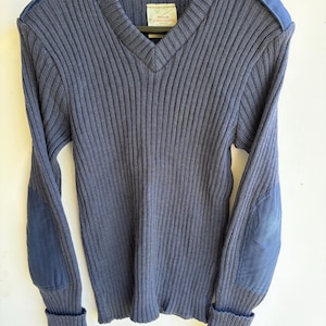 May include: A navy blue V-neck sweater featuring a ribbed knit texture, long sleeves with solid fabric elbow patches, and matching fabric shoulder epaulets with buttons. A white rectangular label is stitched inside the neckline.