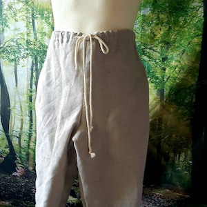 May include: Beige linen pants with a drawstring waist. The pants are displayed on a wooden mannequin. The fabric appears to be lightweight and breathable, suitable for warm weather. The pants have a relaxed fit and a simple design.