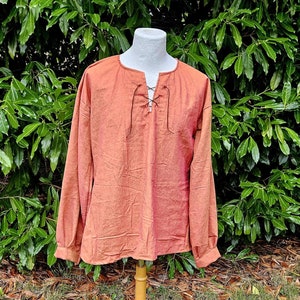 May include: A long-sleeved, rust-colored shirt with a lace-up neckline. The shirt is displayed on a wooden mannequin against a backdrop of green foliage.