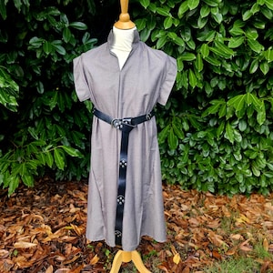 May include: A gray dress with short sleeves and a high collar is displayed on a wooden mannequin. The dress is cinched at the waist with a black belt. The dress is knee-length and has a button-down front.