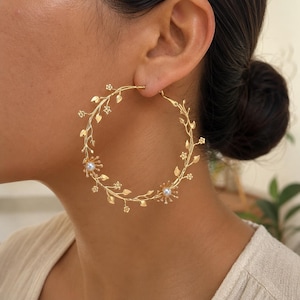 May include: A large gold hoop earring featuring a delicate vine design with small metallic leaves and tiny flowers. Two small white pearls are set within the floral accents, positioned along the circular frame of the earring, creating a detailed botanical appearance.