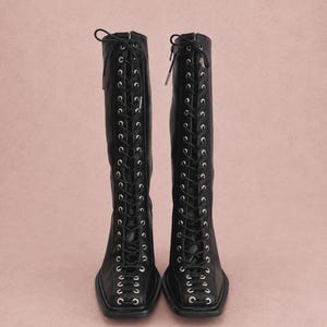 Peut inclure: Une paire de bottes noires arrivant au genou, dotées d'un laçage frontal complet avec des œillets métalliques argentés. Ces bottes présentent une silhouette à bout carré, un talon bloc et des lacets noirs noués en haut, offrant un aspect structuré.