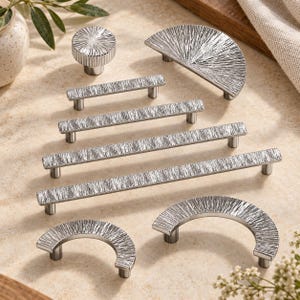 May include: A collection of silver-toned cabinet hardware featuring a textured, radial-line finish. The set includes one round knob, one semi-circular pull, two arched pulls, and four linear bar pulls of varying lengths, all with a metallic sheen.