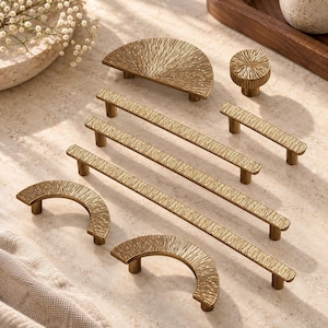 May include: A collection of brass-toned cabinet hardware featuring a textured, sunburst-like pattern. The set includes three long straight bar pulls, two semi-circular arch pulls, one half-circle pull, and one small round knob, all with a metallic finish.