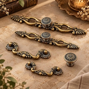 May include: A collection of antique brass-toned metal hardware including two long ornate drawer pulls with central knobs, two smaller curved drop-style drawer pulls, and one round cabinet knob, all featuring intricate floral and leaf-like detailing.