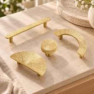 May include: A collection of four gold-toned cabinet hardware pieces featuring a textured, radial pattern. The set includes one long rectangular bar pull, one circular knob, one semi-circular arch pull, and one half-circle pull, all with a metallic finish.