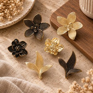 Puede incluir: Un conjunto de seis pomos decorativos de metal con diversas formas de flores y hojas. El set incluye tres pomos con forma de flor en acabados bronce oscuro y dorado, y tres pomos con forma de hoja en tonos metálicos de bronce oscuro y oro.